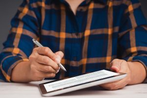 Business professional in plaid shirt using a stylus to sign and annotate a digital document on a tablet, performing detailed contract analysis for review and approval.