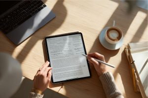 Business professional reviewing digital charts and analytics on a tablet at a sunlit workspace, with coffee and documents nearby, representing the analysis and preparation phase before finalizing a sales contract.